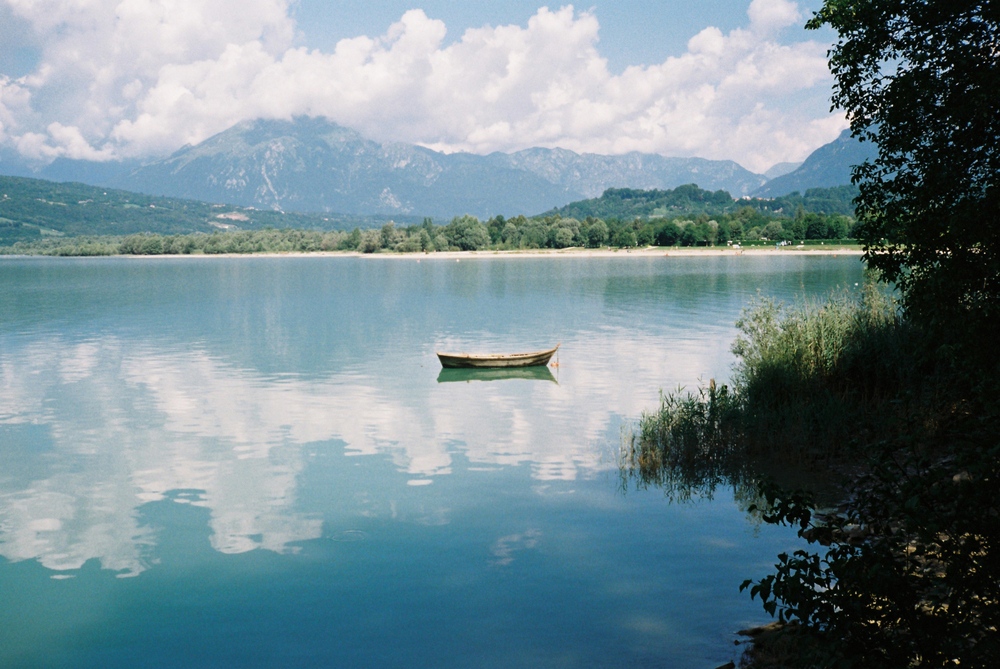 A small wooden boat floating on a calm lake with mountains and clouds reflected in the water.